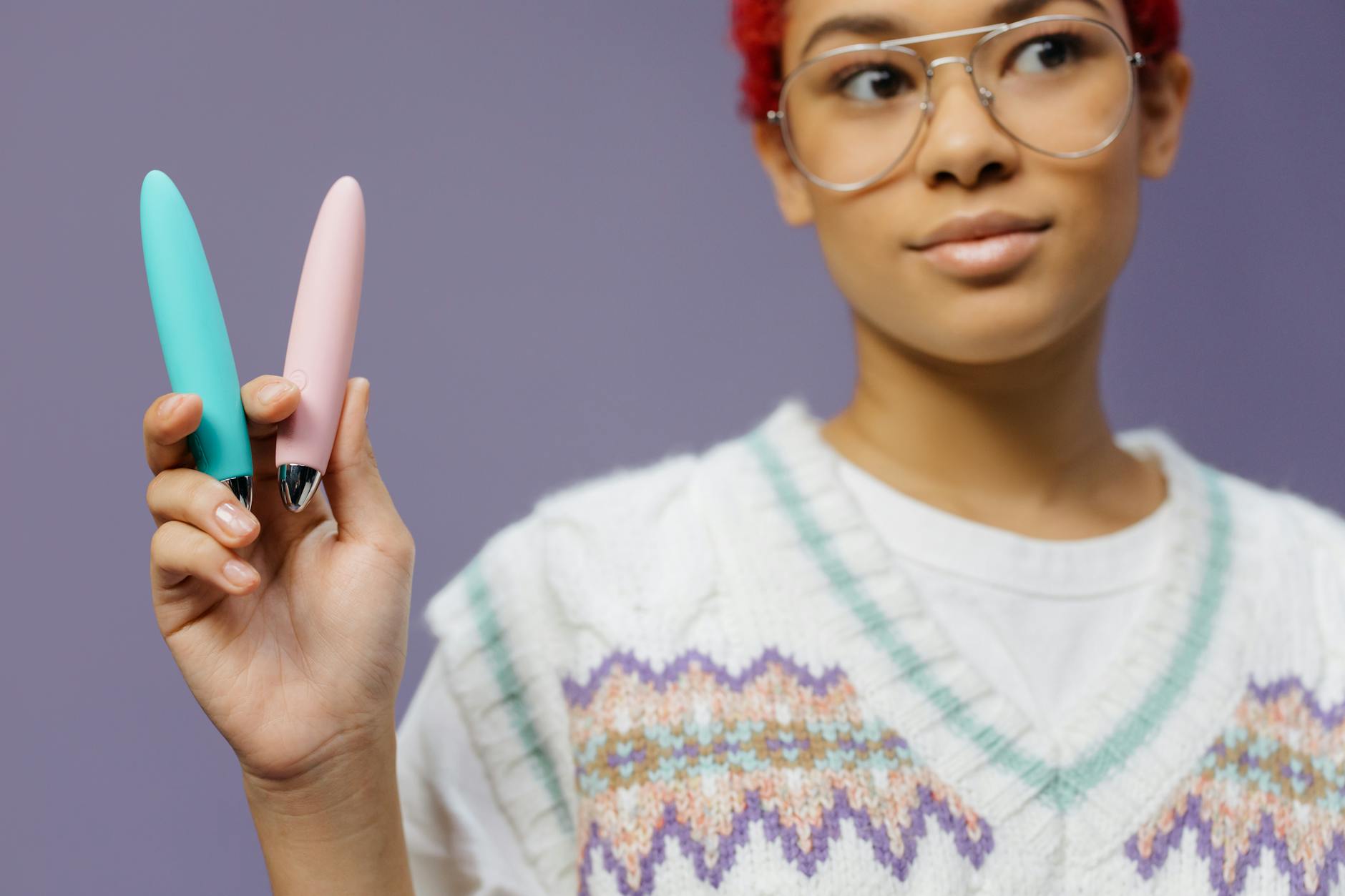 Close-up of two hands holding colorful vibrators against a purple background